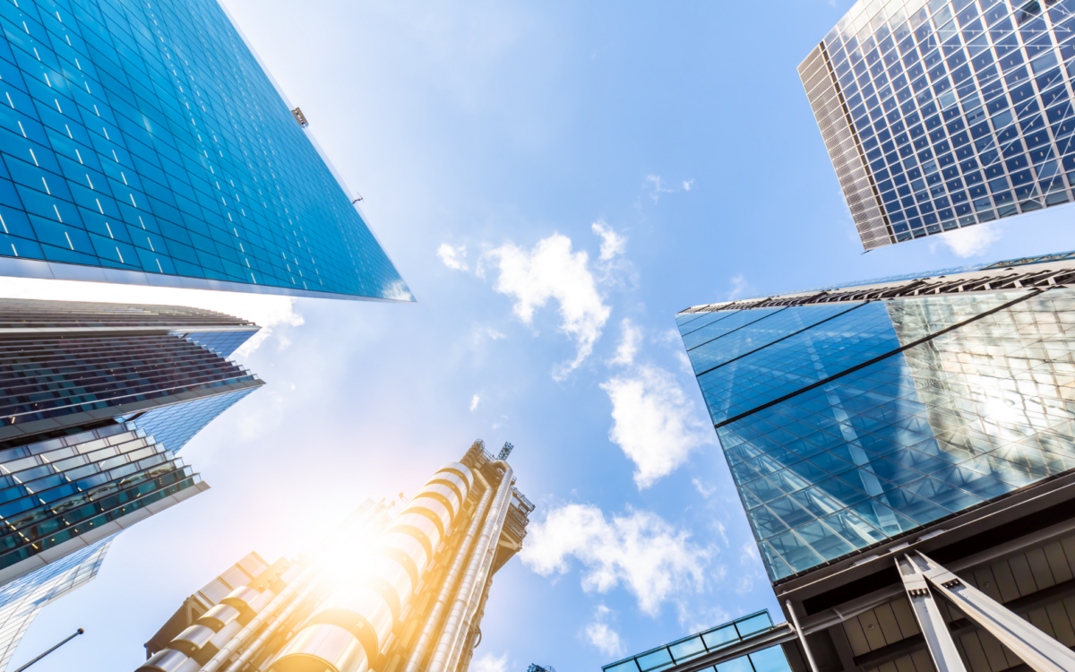 Upward view of modern skyscrapers in London's financial district, including iconic buildings associated with the reinsurance and insurance industries, reflecting contemporary architecture and urban development against a bright blue sky with scattered clouds.